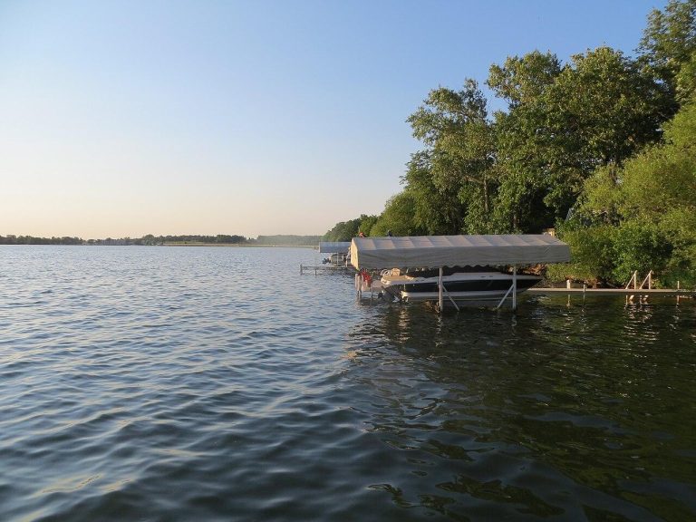 A tranquil lakeside scene at Detroit Lakes, featuring a wooden dock shaded under a gray canopy, gently bobbing on rippling waters. Tall green trees line the distant shore under a clear blue sky, reflecting a serene golden glow of late afternoon.