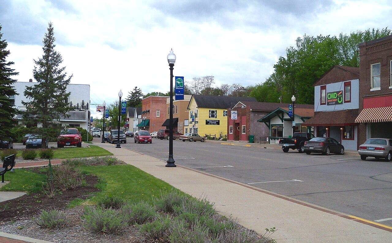 A calm street scene in Watertown featuring charming buildings in subtle hues of yellow and red. Lush green grass and low shrubs line the sidewalk, while classic lampposts stand tall with banners, enhancing the quaint atmosphere of this small-town setting.