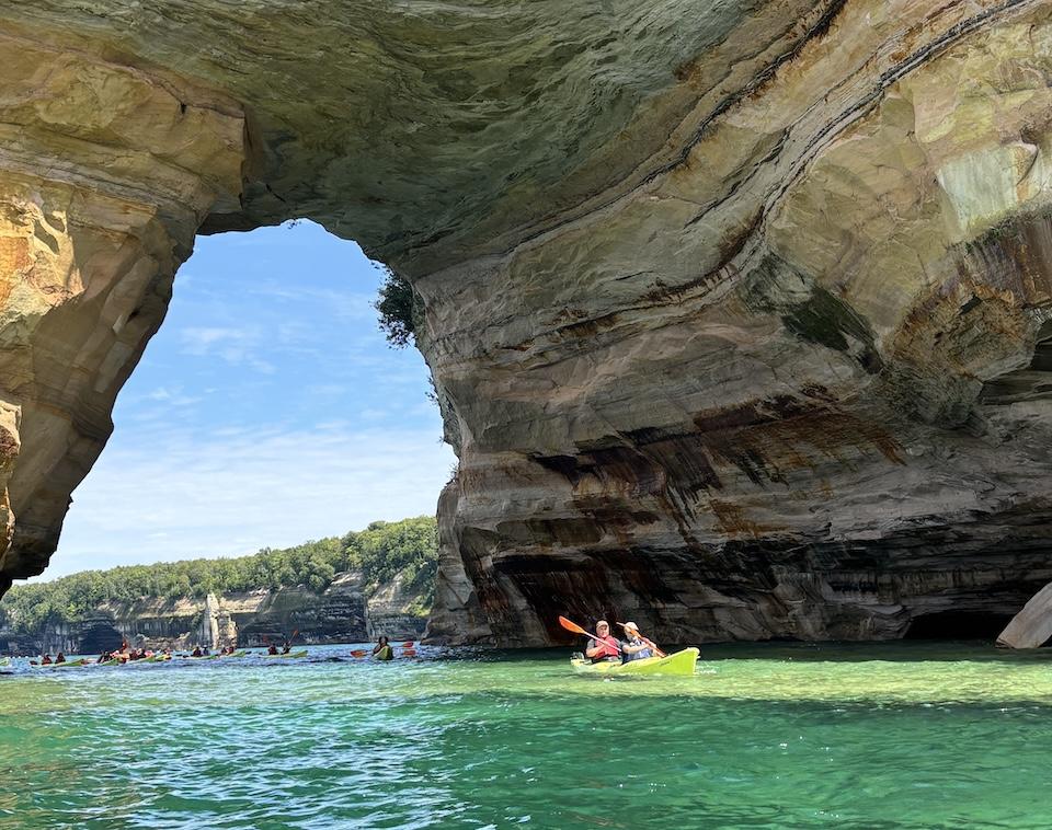 Two guys on a double kayak paddling under a rock formation.